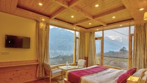 A room with a large bed, a desk with a chair, a TV mounted on the wall, and large windows offering a mountain view. - Sparklyn View (Manali)