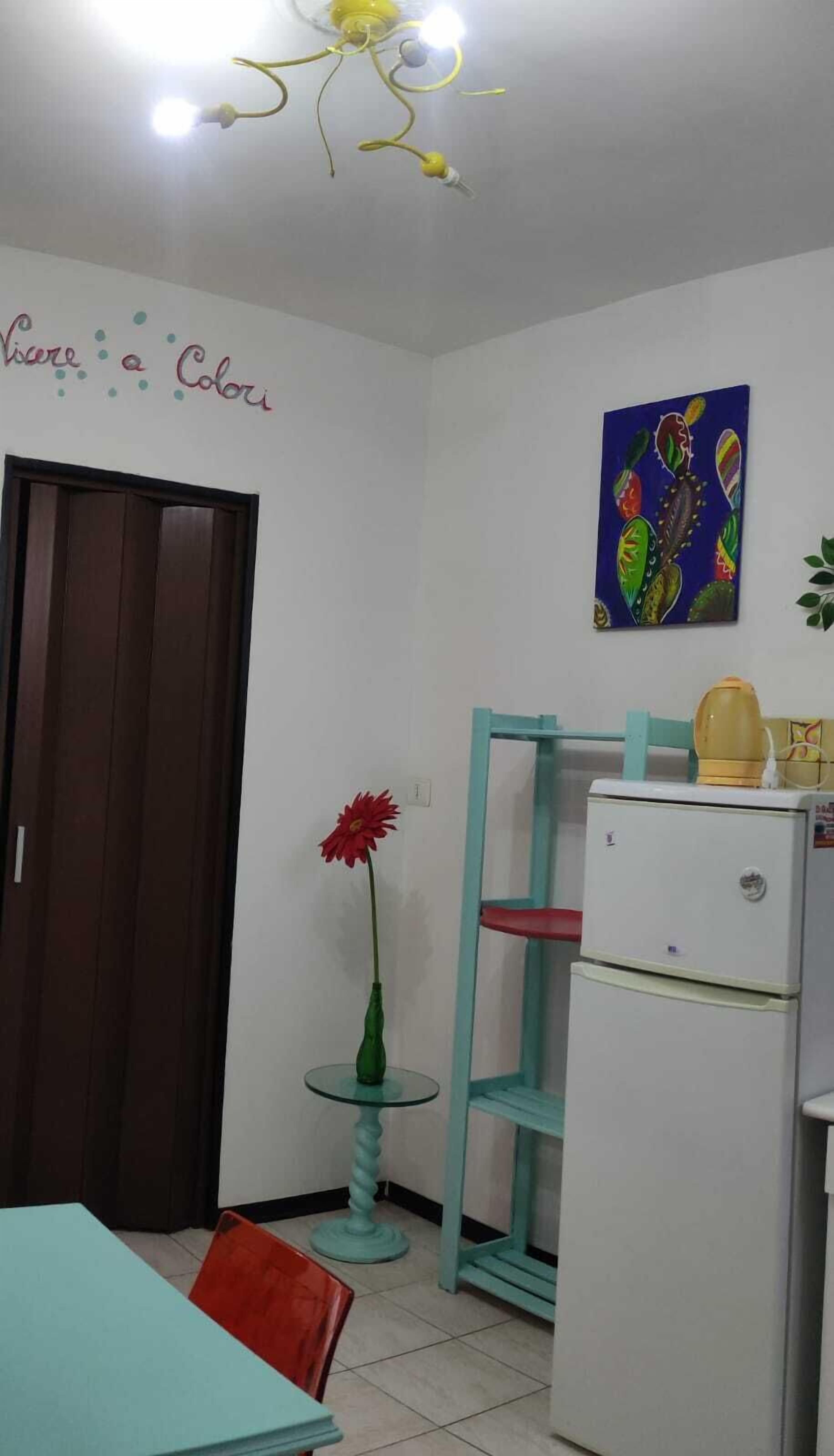 A small, well-lit room with a refrigerator, a table with chairs, a small plant, and a painting on the wall.