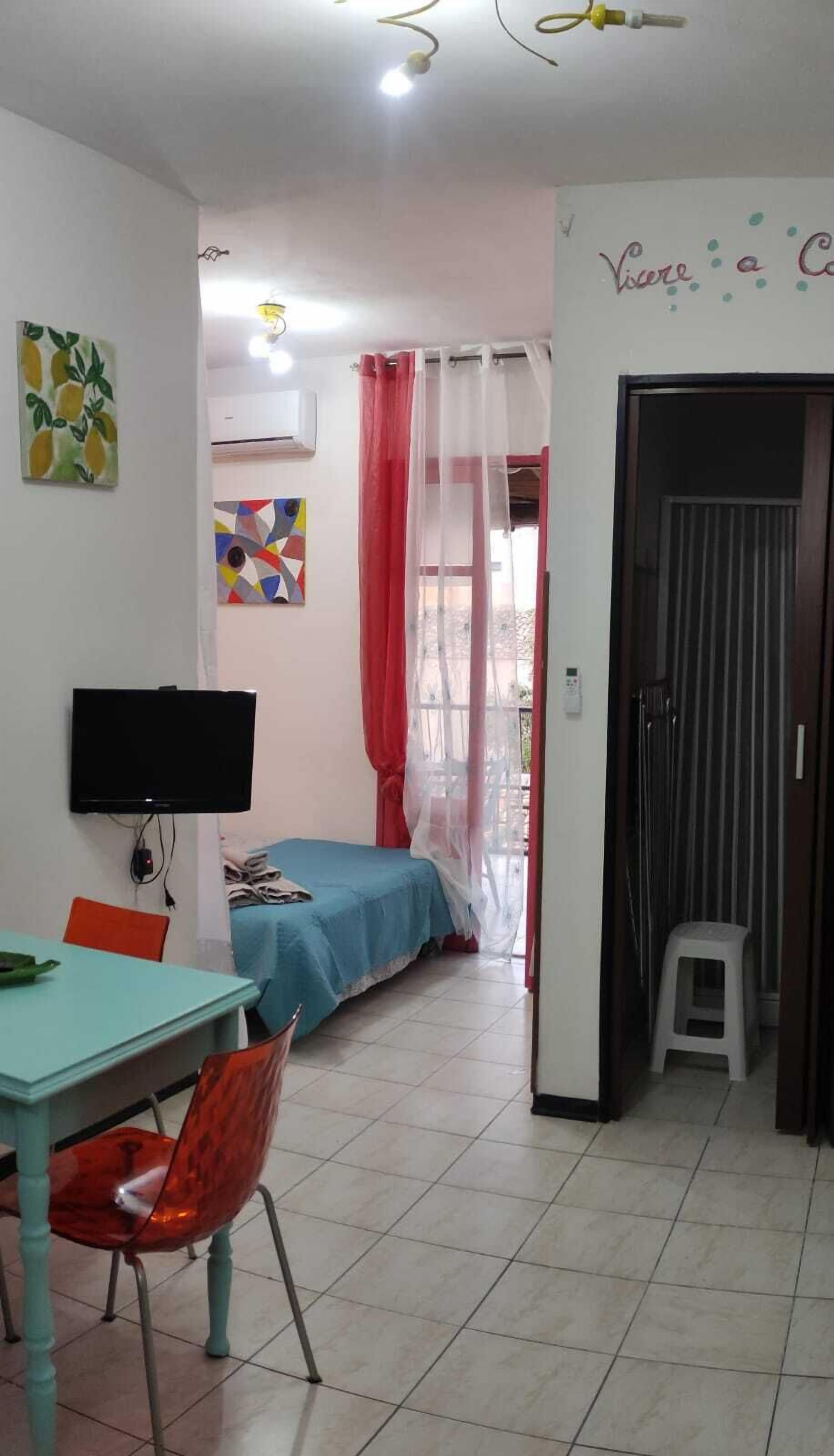 A room with a bed, a TV, a table, and chairs. There is a door leading to another room.