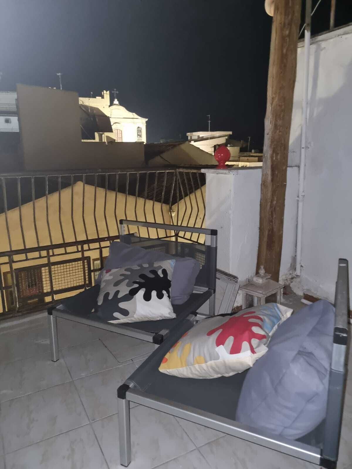 En balkon med gelænder, en seng med puder, og bygninger i baggrunden.