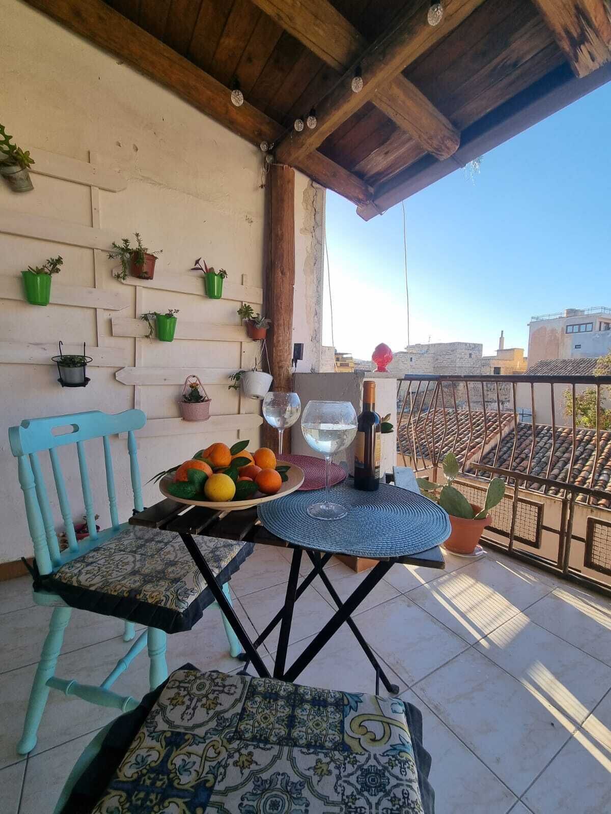 Un balcón con techo de madera, una mesa con mantel tejido, una silla y varias plantas en macetas en la pared.