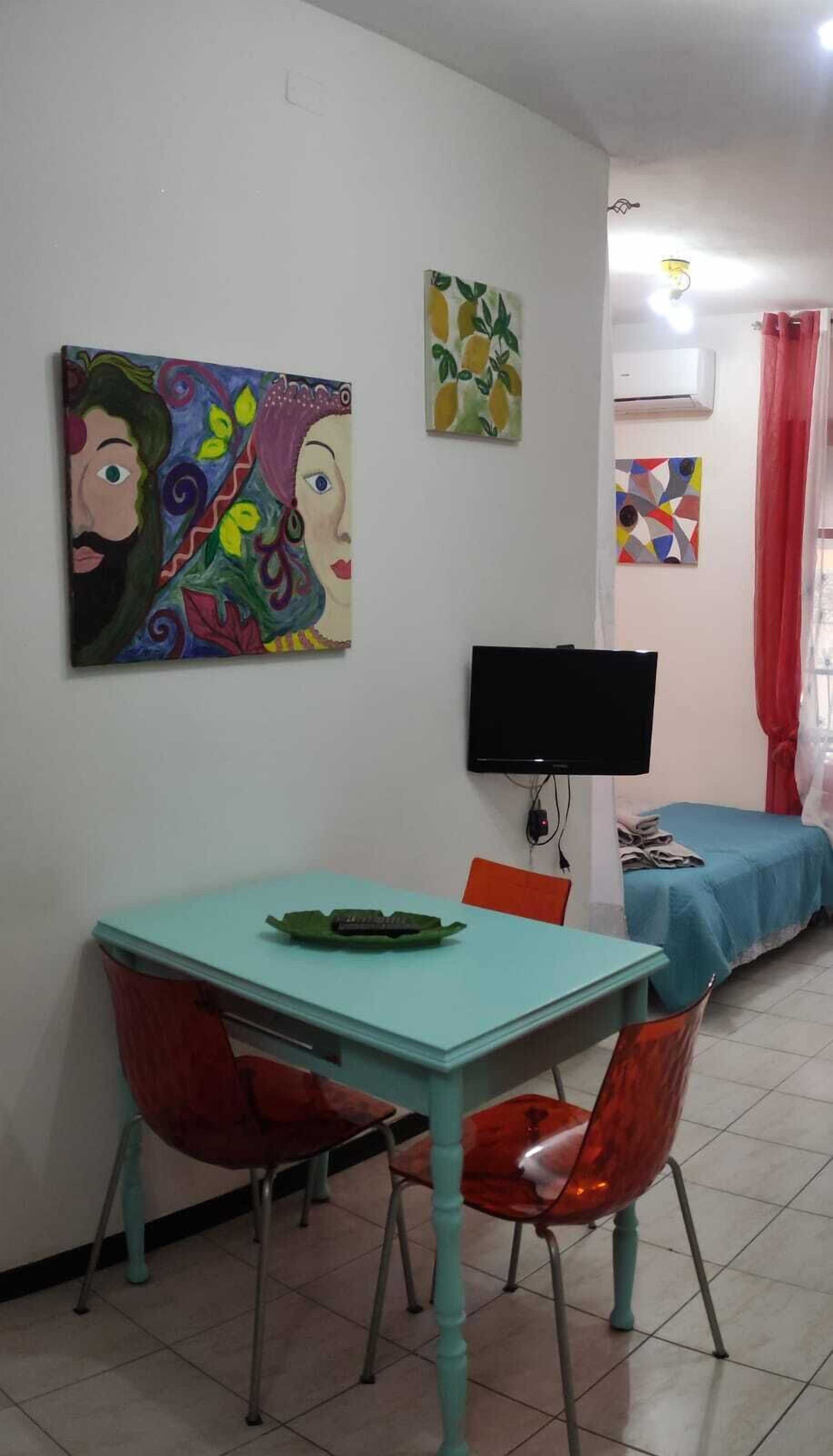 Un comedor con una mesa turquesa y sillas rojas, un televisor de pantalla plana y obras de arte en las paredes.