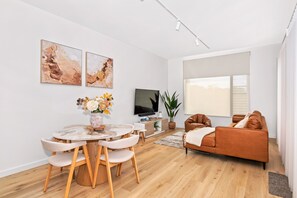 Un salon moderne avec une table de salle à manger ronde, deux chaises, un canapé, un téléviseur et des œuvres d’art accrochées au mur.