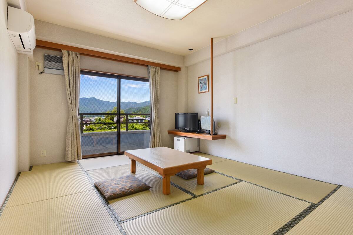 A room with tatami flooring, a wooden table, a TV on a shelf, and a view of mountains through a sliding door.