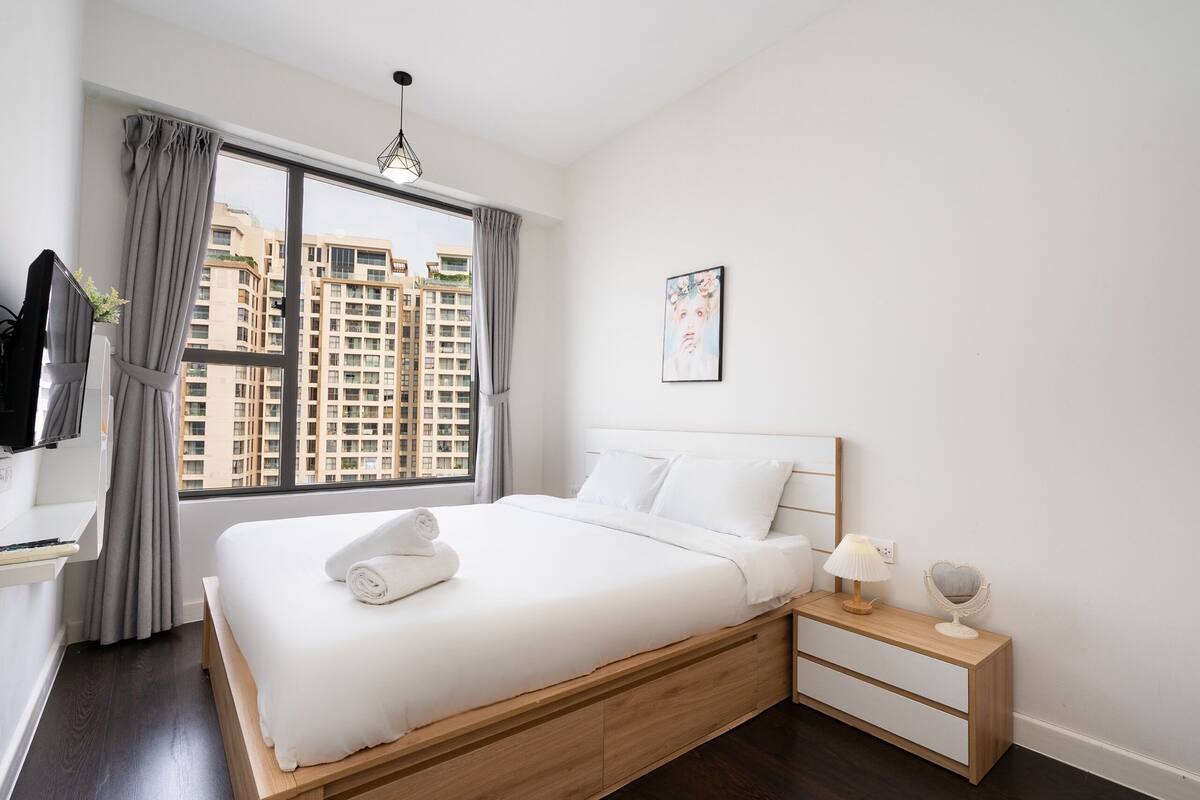 A modern hotel room with a large window, a neatly made bed, a bedside table with a lamp, and a wall-mounted TV.