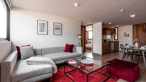 Ein modernes Wohnzimmer mit Sofa, Couchtisch und rotem Teppich. Es gibt einen Essbereich mit Tisch und Stühlen. Im Hintergrund ist ein großes Fenster und eine Küche zu sehen.