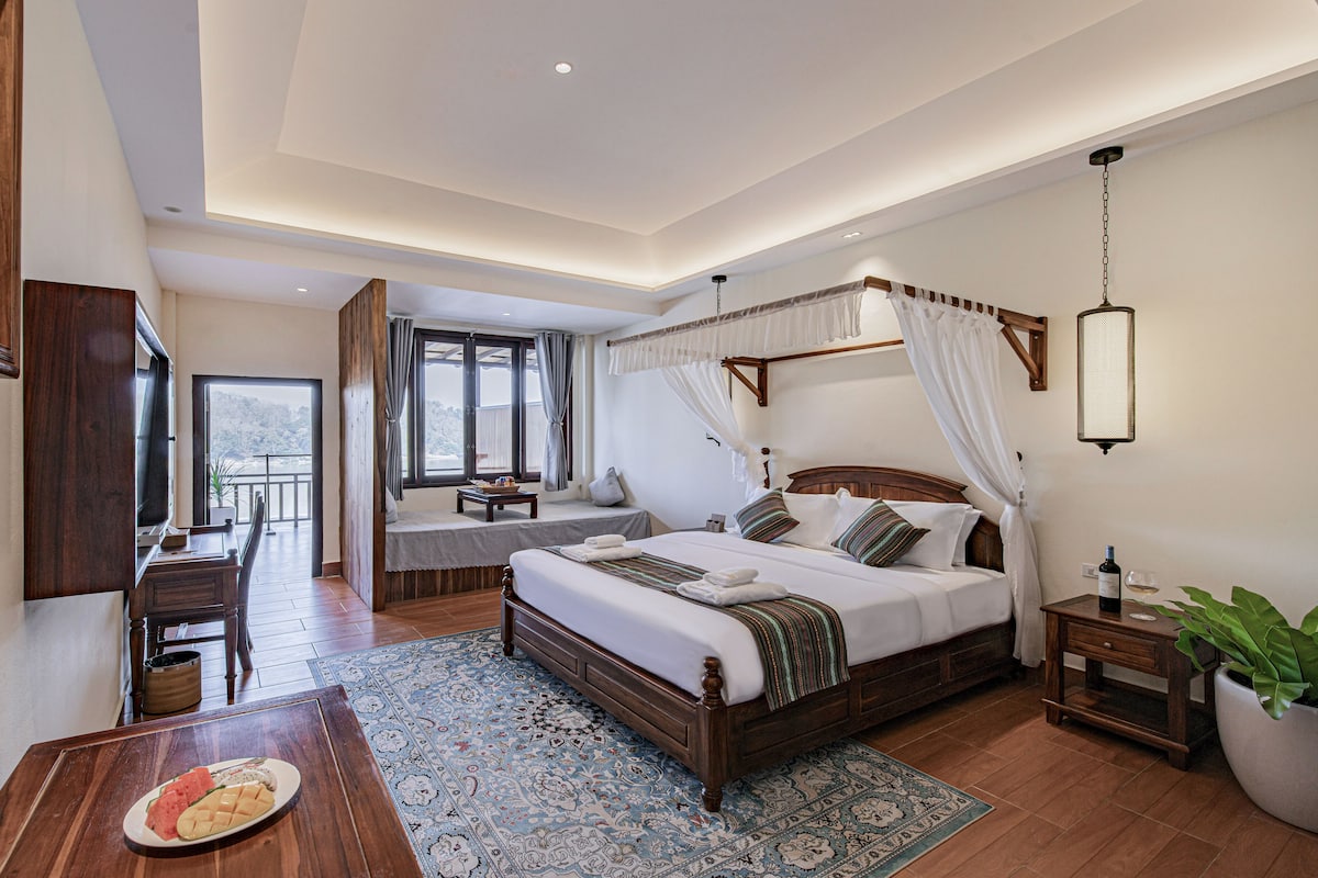 A spacious bedroom with a large bed, a bedside table, a nightstand, a potted plant, and a view of the outdoors.
