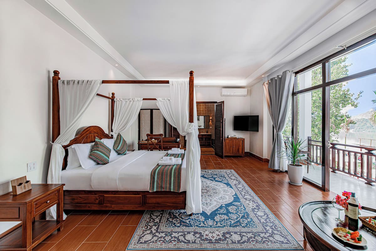 A spacious bedroom with a four-poster bed, a large window with a view, a sitting area with a table and chairs, and a bathroom with a sink and mirror.