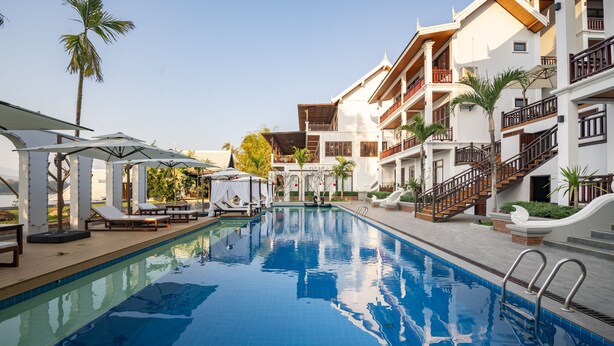 A swimming pool with lounge chairs and umbrellas, surrounded by multi-story buildings with balconies and stairs. - Relaxful Hotel (Luang Prabang)