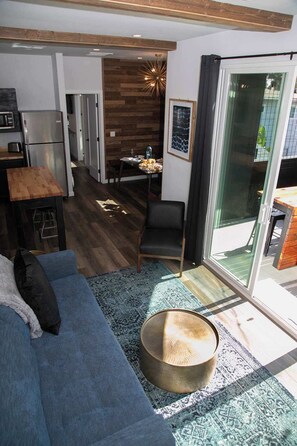 A modern living room with a sofa, a dining area with a table and chairs, a kitchen with appliances, and a large sliding glass door leading to an outdoor area.