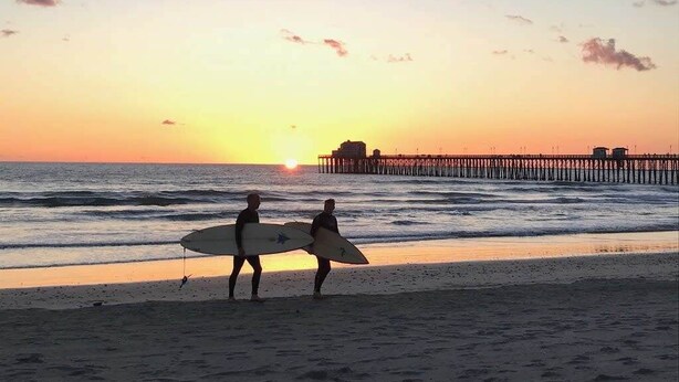 A beach scene with two people carrying surfboards, a pier in the background, and the sun setting over the ocean. - Industrial-Inspired 2-Story California Beach House (Oceanside)