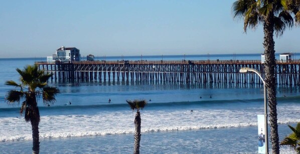 A pier extending into the ocean with a building at the end. Palm trees are visible in the foreground.