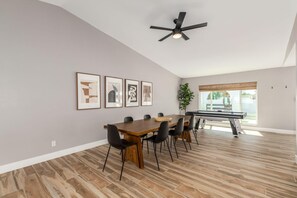 A spacious room with a dining table, chairs, a ceiling fan, framed artwork on the wall, and a large window with a view.
