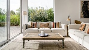 A modern living room with a large window, a wooden coffee table, and two white sofas with patterned cushions. - Eco-Luxe Villa Laranjeiras Comporta - Pool & Golf (comporta)