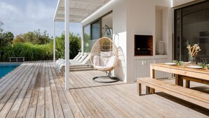 A wooden deck with a white canopy, a wicker chair, a wooden table with plates and glasses, and a large window overlooking greenery. - Eco-Luxe Villa Laranjeiras Comporta - Pool & Golf (comporta)