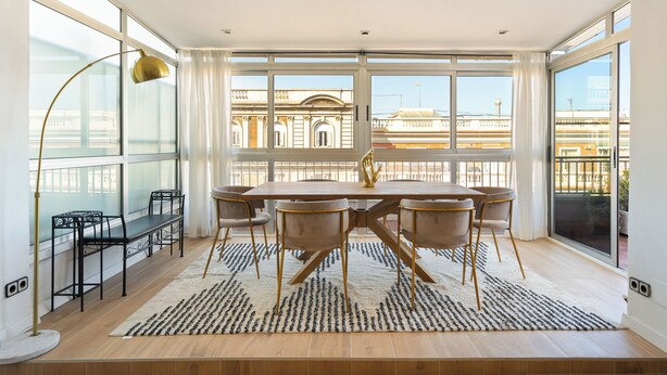 A dining area with a large table, chairs, a rug, and a view of buildings through large windows.