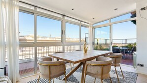 A spacious room with a large wooden table and four chairs, large windows with a view of a cityscape, and a balcony with outdoor furniture.