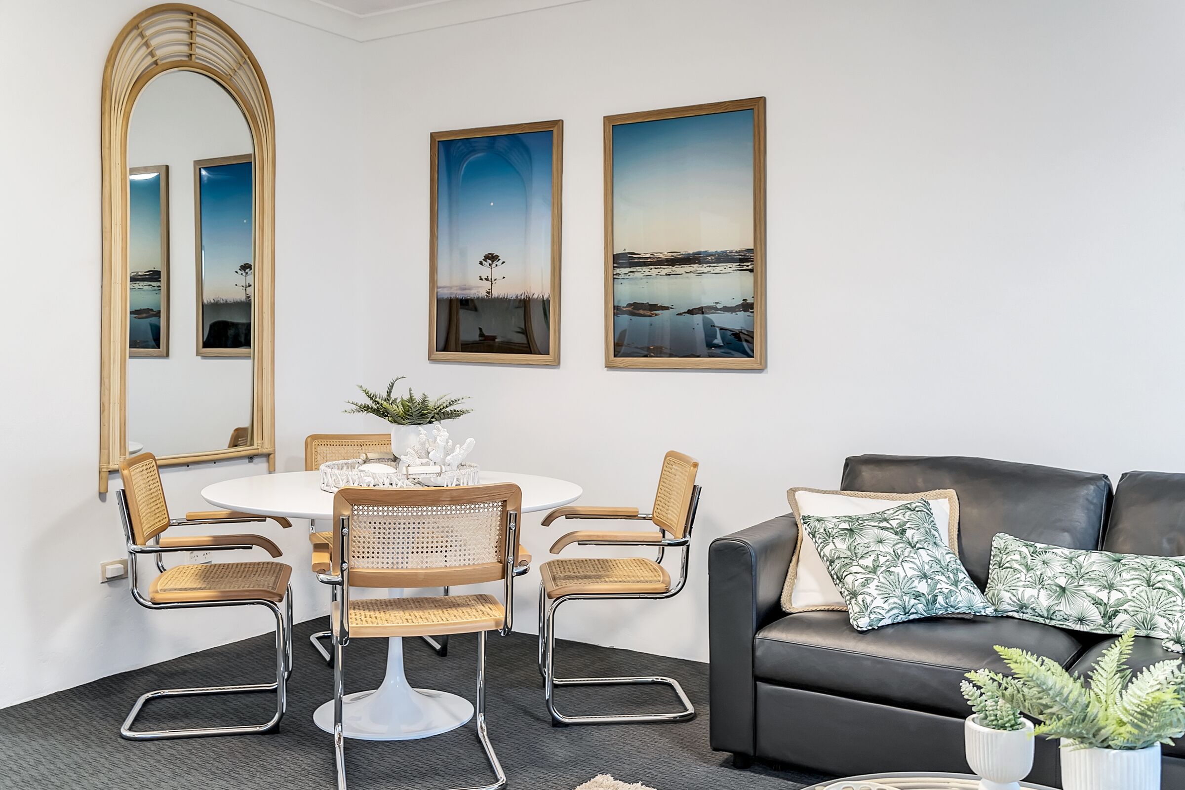A modern living room with a round dining table, four wicker chairs, a black leather sofa, and three framed landscape paintings.