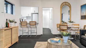 Uma sala de estar moderna com um sofá, poltronas, uma mesa de centro e uma área de jantar com mesa e cadeiras. Uma cozinha é visível ao fundo.