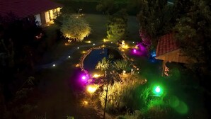 A well-lit garden with a circular pond, surrounded by trees and a small building with a red roof. - Ferienhaus am Glück (Bad Zwischenahn)