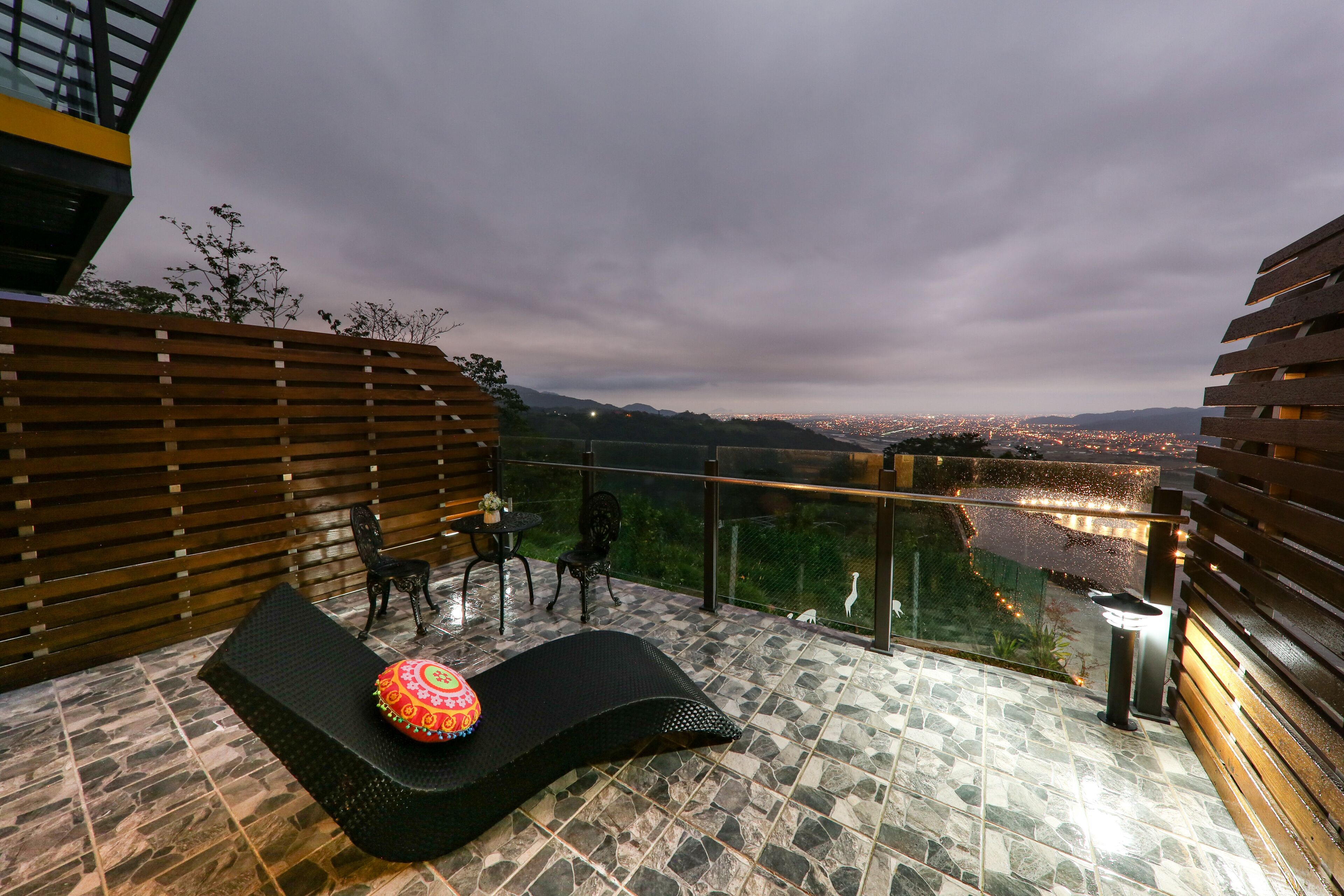 Un balcón con una silla lounge negra, una mesita y sillas. El balcón ofrece vistas a un paisaje urbano nocturno.