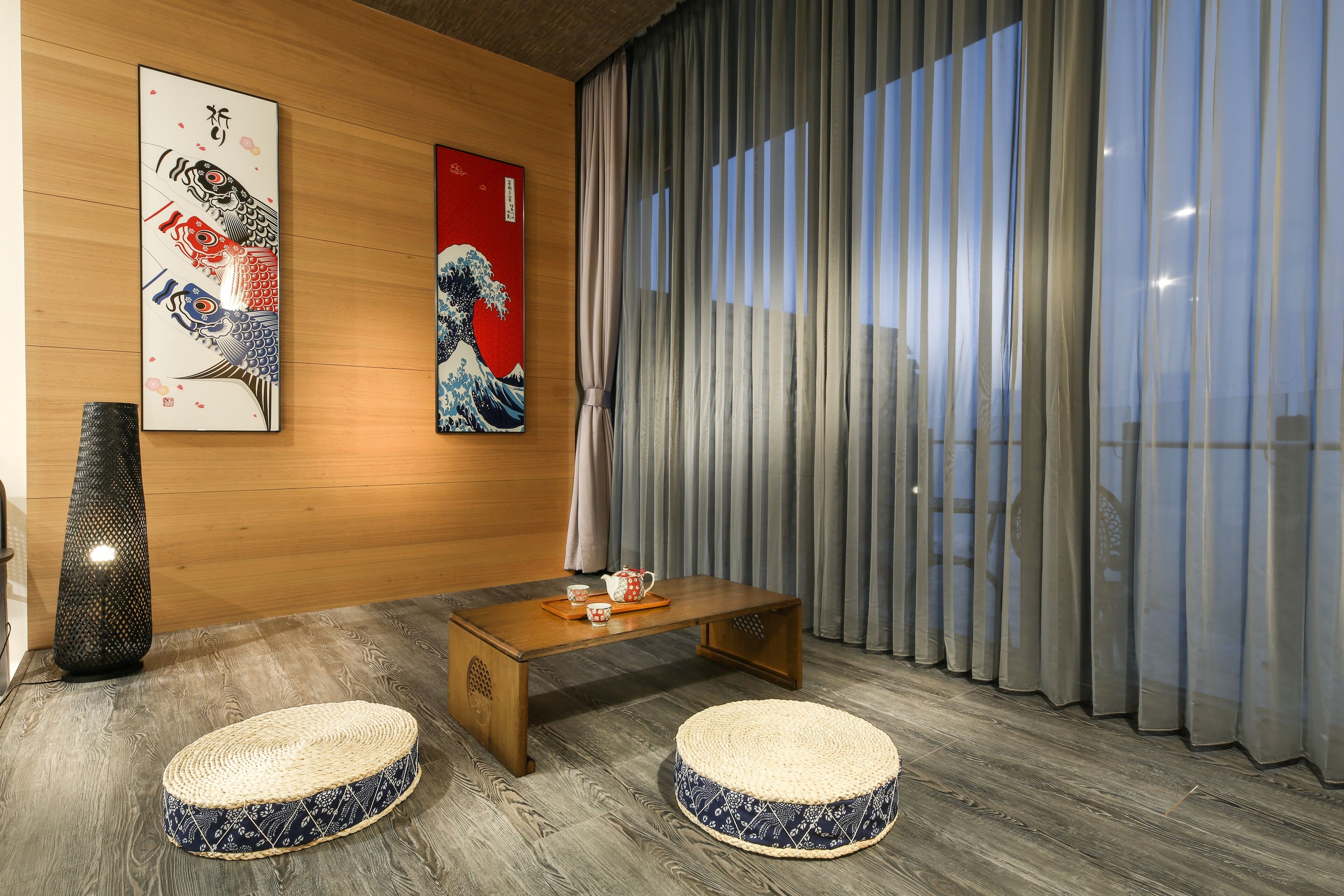 Una habitación con piso de madera, una mesa de centro de madera, dos otomanas tejidas, un jarrón alto y dos obras de arte enmarcadas en la pared.