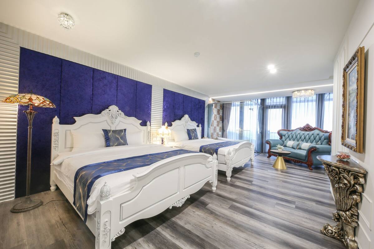 Un dormitorio con dos camas blancas, una pared azul decorativa, una araña y un sofá azul.