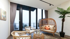 Ein modernes Wohnzimmer mit Rattanmöbeln, einer großen Kübelpalme und Blick auf die Stadt durch große Fenster.