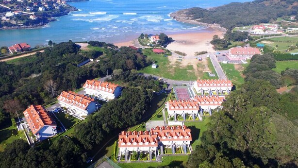 An aerial view of a coastal resort with multiple buildings, a beach, and a mountainous backdrop.