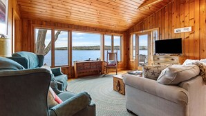 A wooden cabin interior with a sofa, a chair, a television, and a view of a lake. - Tide's Edge-New Oceanfront! (Surry)