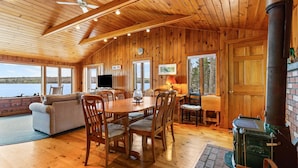 A wooden cabin interior with a dining table, chairs, a sofa, a fireplace, and large windows overlooking a lake. - Tide's Edge-New Oceanfront! (Surry)