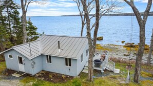 A small, light blue cabin with a metal roof, situated by a rocky shoreline with a view of a large body of water. - Tide's Edge-New Oceanfront! (Surry)