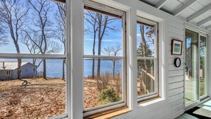 A room with large windows overlooking a body of water, a wooden deck, and a view of trees. - Seaside! New-Oceanfront! (Surry)