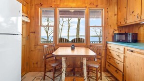 A wooden cabin kitchen with a table and chairs, a refrigerator, a microwave, and a view of trees and water through large windows. - Seaside! New-Oceanfront! (Surry)