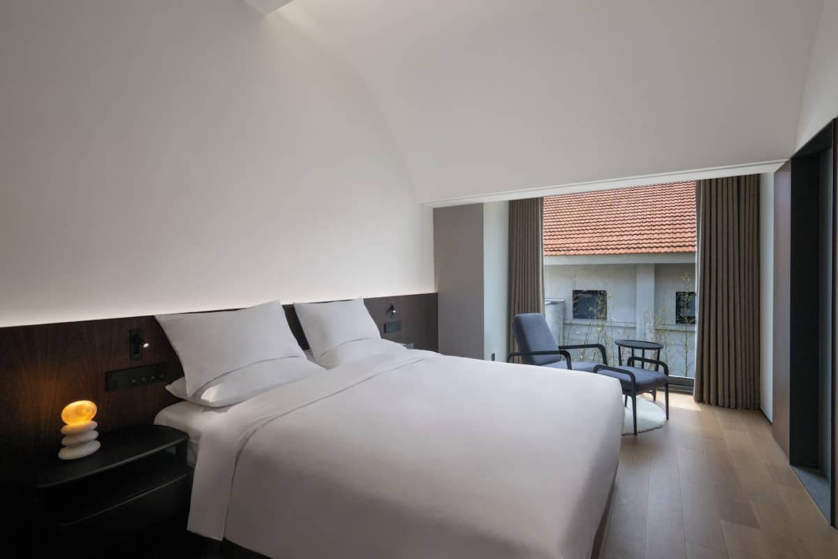 A modern hotel room with a large bed, a nightstand, a desk lamp, and a view of the outdoor terrace.