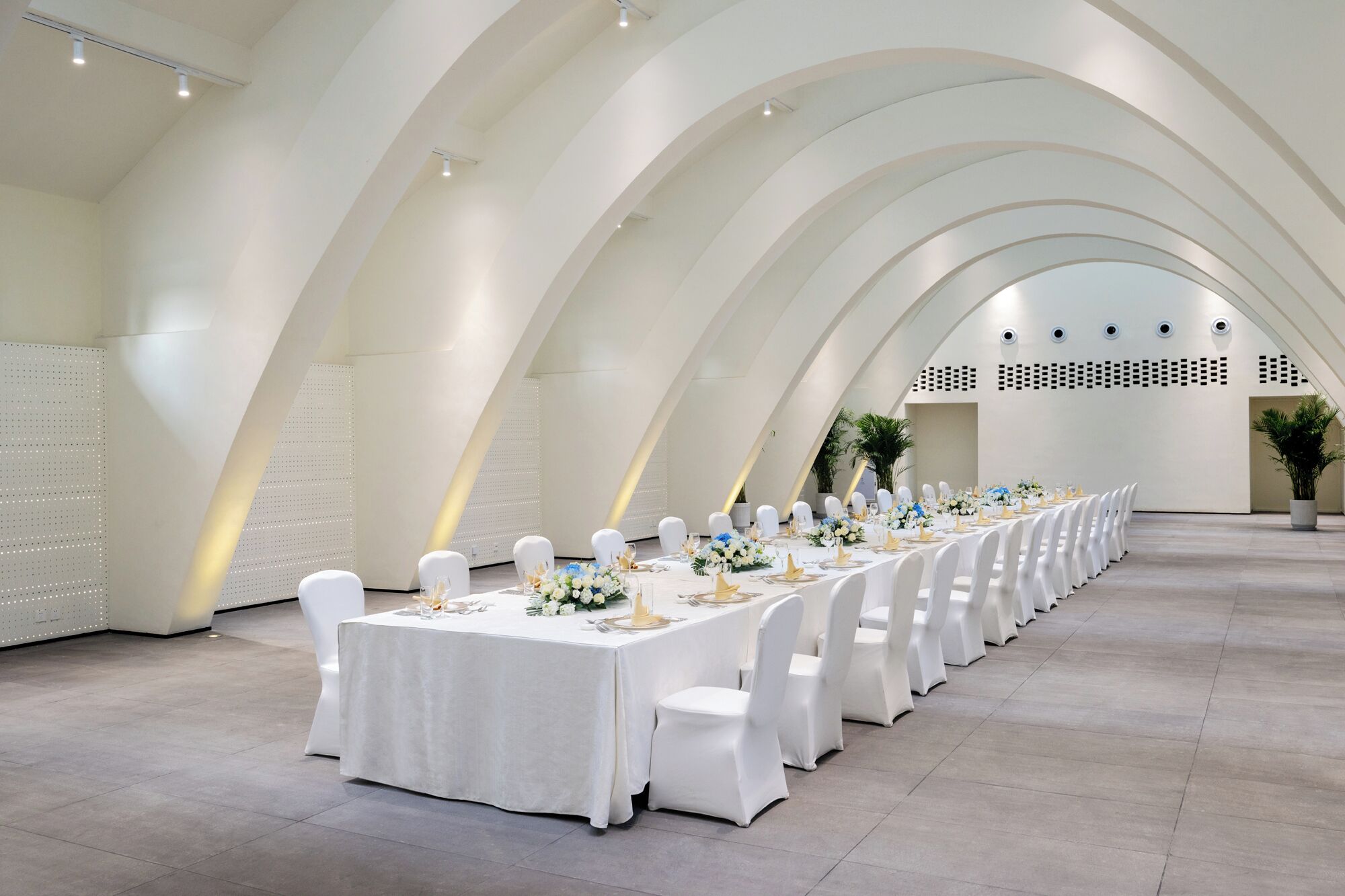 Una larga mesa de banquete dispuesta con manteles blancos, sillas y centros de mesa florales en una sala moderna con techo abovedado.
