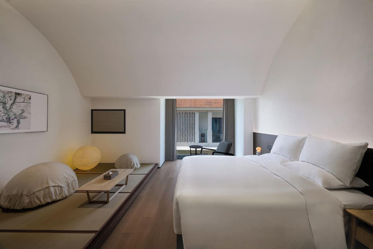 A modern hotel room with a large bed, a small table, and a chair. There is a window with a view of the outside.