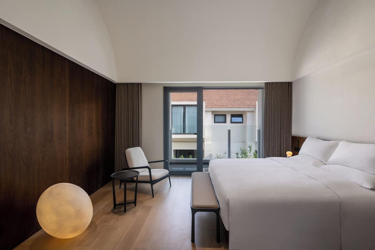 A modern hotel room with a large bed, a round table, a chair, and a bench.