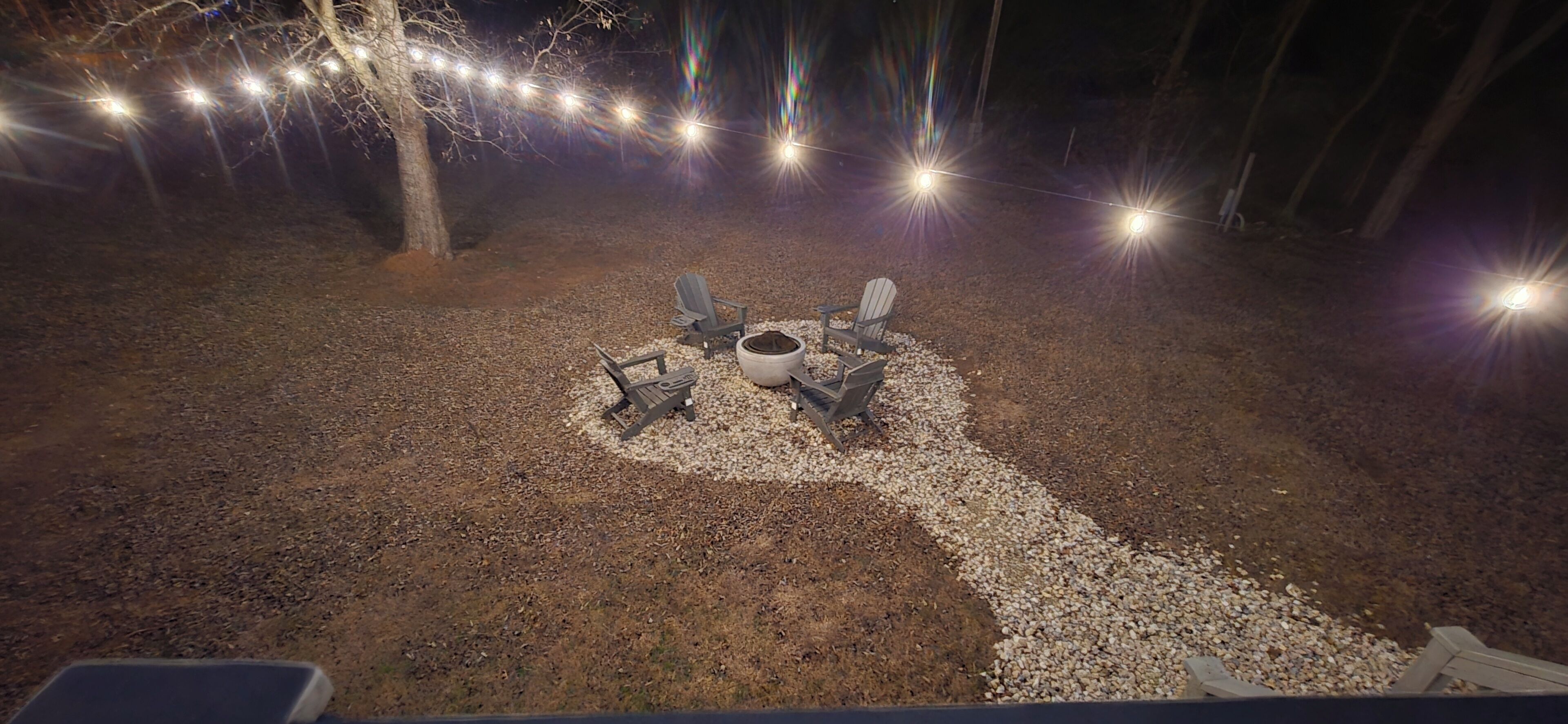 An outdoor seating area with chairs and a fire pit, surrounded by gravel and trees, under string lights.