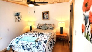 A bedroom with a bed, two bedside lamps, a ceiling fan, and a floral painting on the wall.