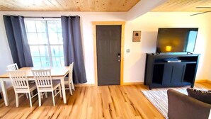 A room with a wooden floor, a dining table with chairs, a dark door, and a TV on a stand.
