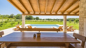 A wooden table with benches under a pergola, overlooking a swimming pool and lush greenery. - Luxury and Stylish Villa Campagna with sauna, private pool and beautiful garden (Pula)