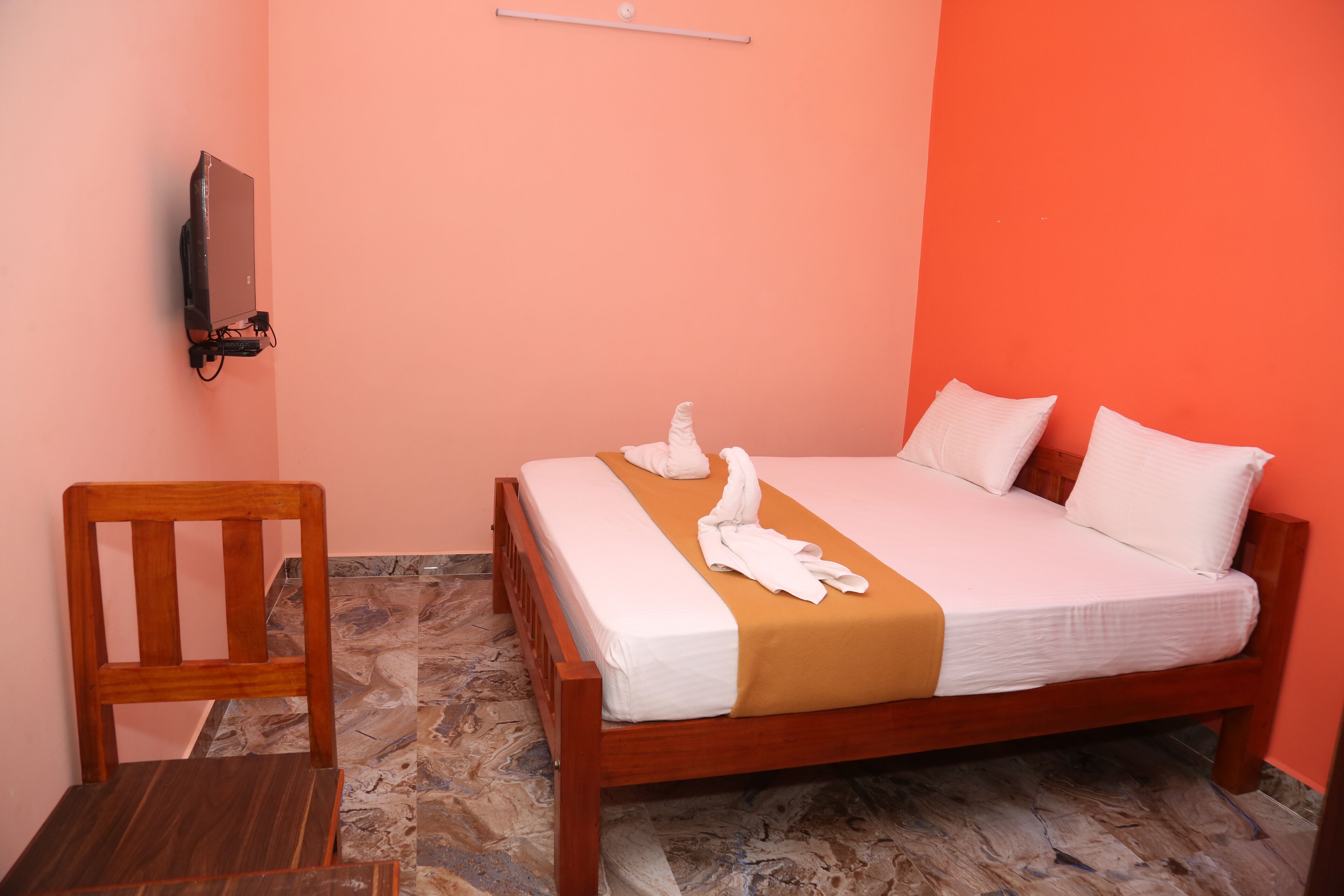A neatly made bed with white and orange bedding, a wooden chair, and a flat-screen TV mounted on the wall.