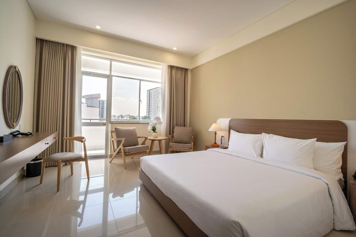 A hotel room with a large bed, a desk, two chairs, a small table, and a view of the city through the window.