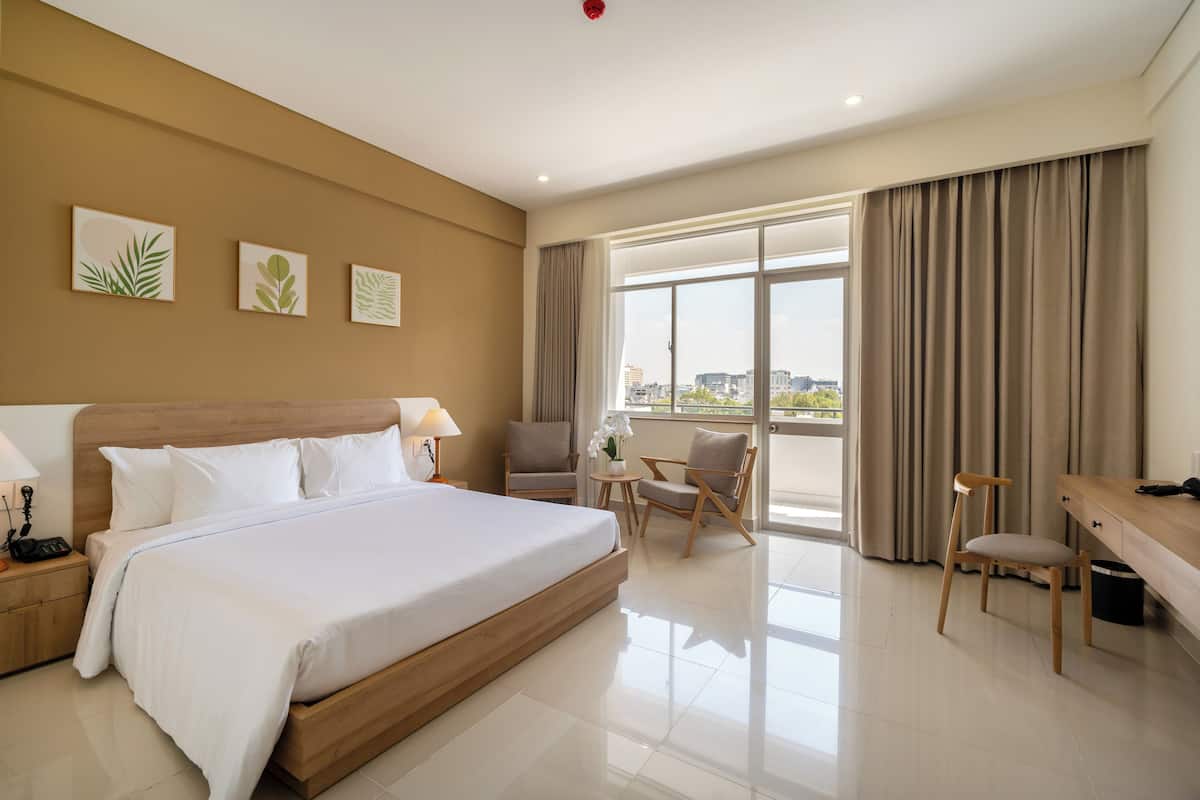 A hotel room with a large bed, a desk, two chairs, a small table with a vase of flowers, and a view of the city through large windows.