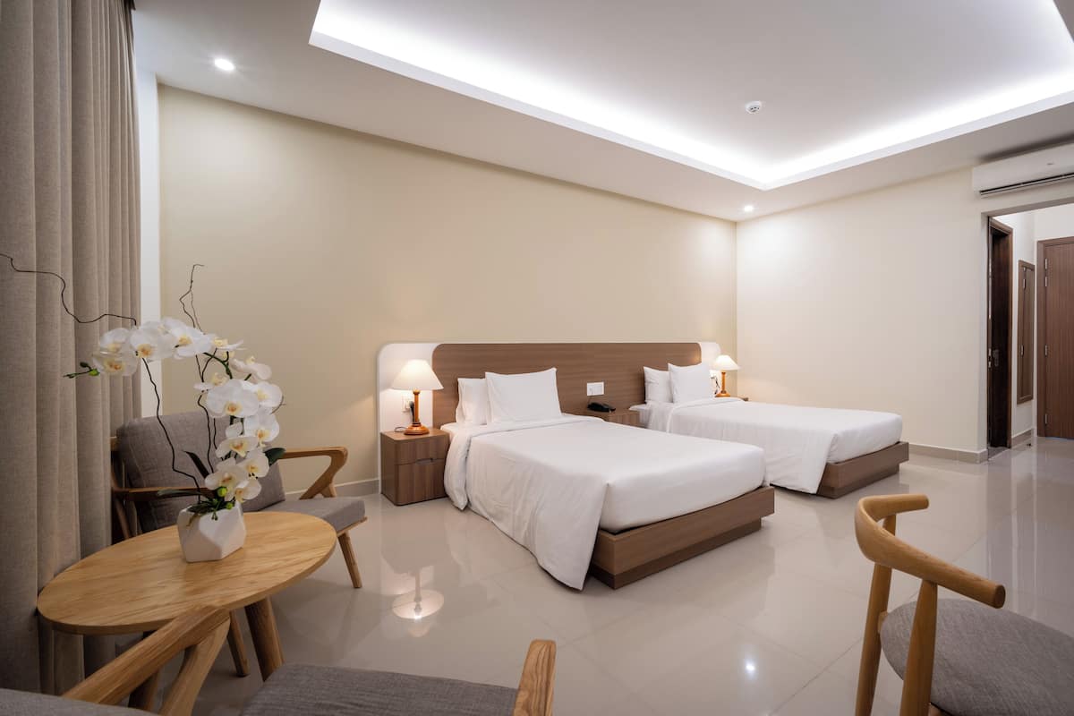 A modern hotel room with two beds, a sitting area with a table and chairs, and a vase of white flowers.