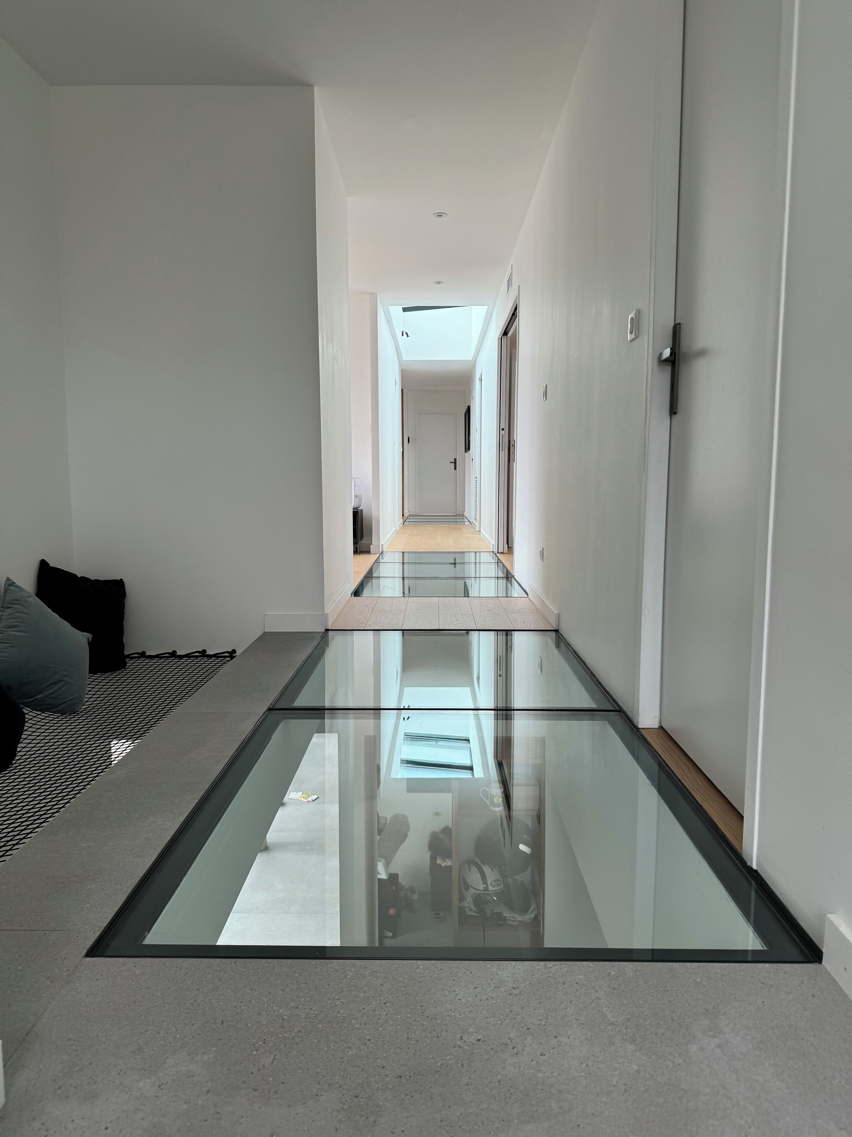 Un intérieur moderne avec un plancher en verre donnant sur un couloir, des murs blancs et une porte sur la droite.