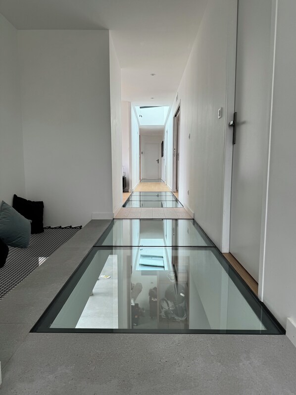 Un intérieur moderne avec un plancher en verre donnant sur un couloir, des murs blancs et une porte sur la droite.