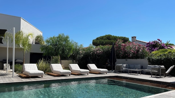 A pool area with four white lounge chairs, a wooden deck, and a white building in the background. - Luxury villa heated pool jacuzzi sauna (Pérols)
