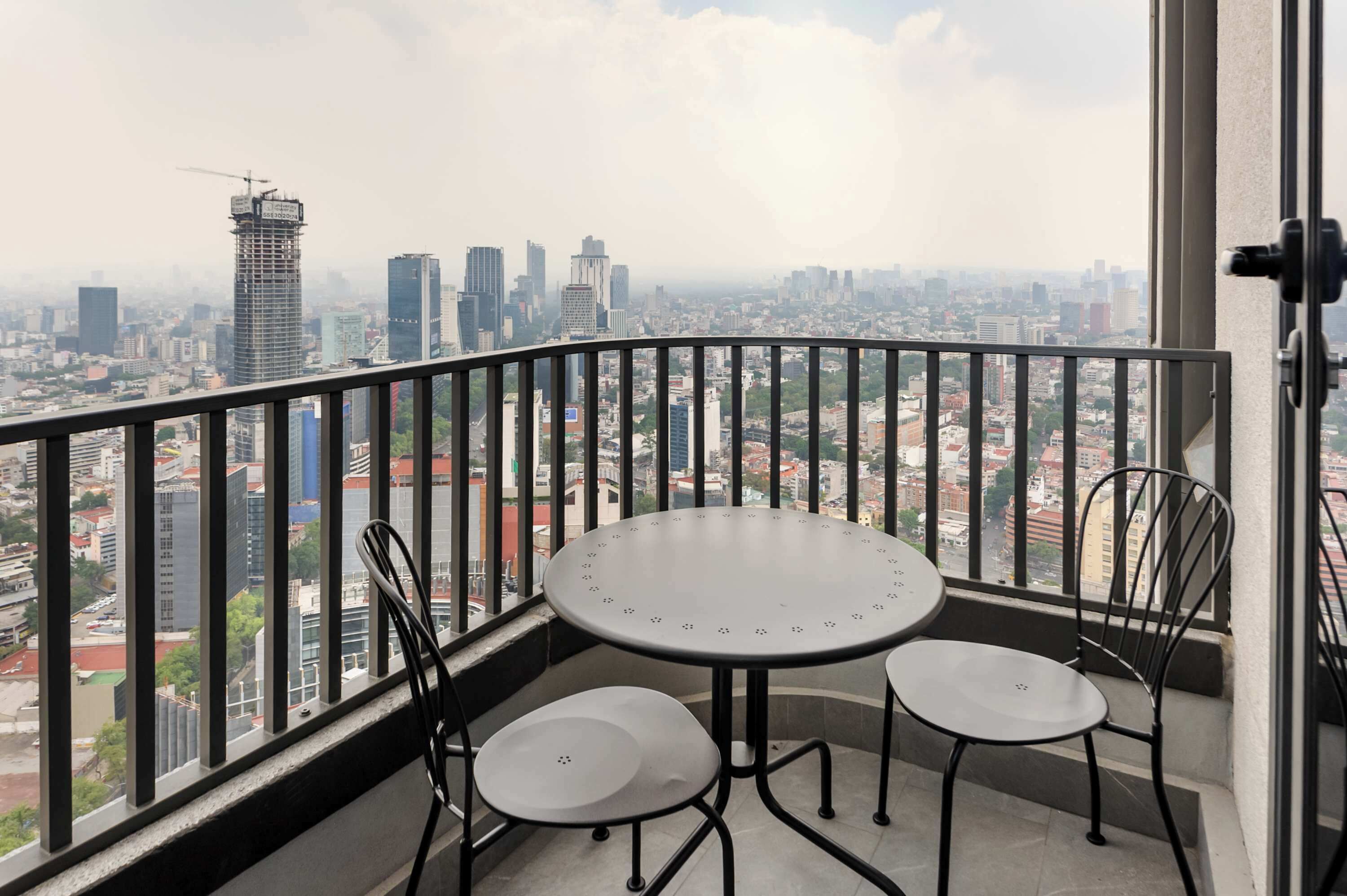A balcony with a round table and two chairs, overlooking a cityscape with various buildings and a construction crane.